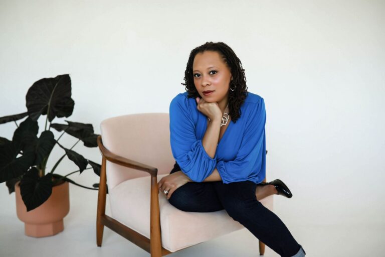 A person is seated in a light pink upholstered chair with wooden armrests, wearing a blue long-sleeved top, dark jeans, and black shoes. One hand rests on the armrest, displaying a ring, while the other hand supports their head. A potted plant with large, dark green leaves is positioned to the left in a terracotta pot. The background is a plain white wall, creating a minimalist setting.