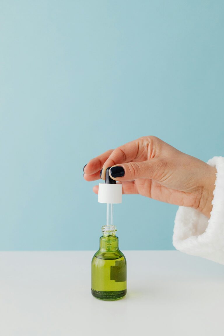 A hand with black-painted nails holds the dropper cap of a green glass bottle containing a yellowish liquid. The bottle is placed on a white surface against a light blue background.