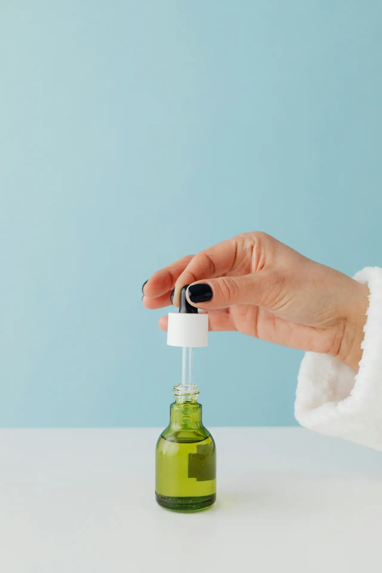 A hand with black-painted nails holds the dropper cap of a green glass bottle containing a yellowish liquid. The bottle is placed on a white surface against a light blue background.