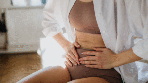 A person sits on a bed in a room with wooden flooring and white walls, illuminated by soft natural light. They are wearing a beige sports bra, matching high-waisted leggings, and an open white button-up shirt. Their hands are placed on their abdomen, with fingers interlocked.
