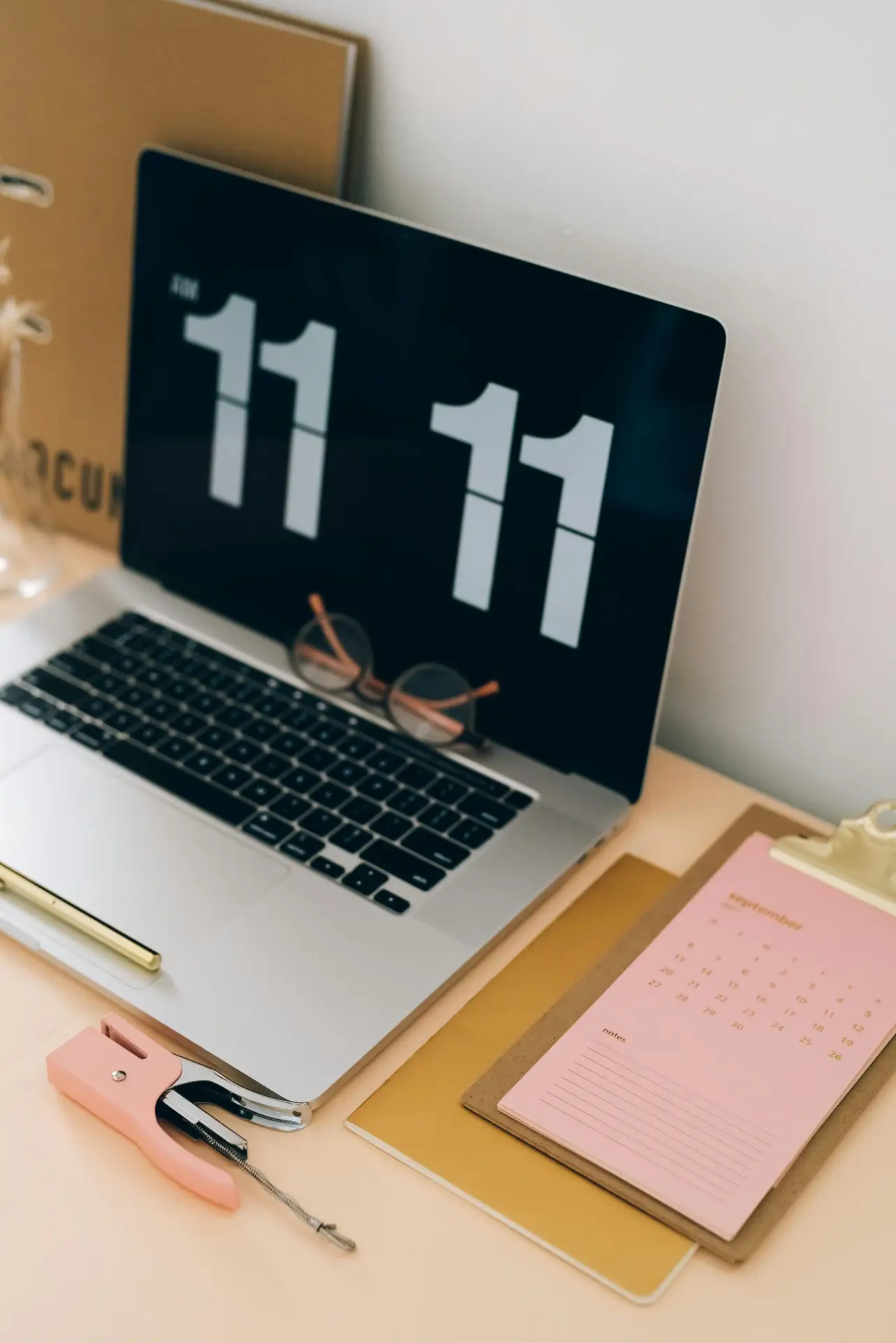 A laptop displaying "11:11" on the screen, a pair of glasses, a pink stapler, a pen, and a calendar dated September on a desk. A notebook is partially visible in the background.