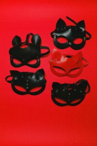 Five leather masks on a red background. Four are black, one is red. All have pointed ears, metal studs, straps, and eye and nose cutouts.