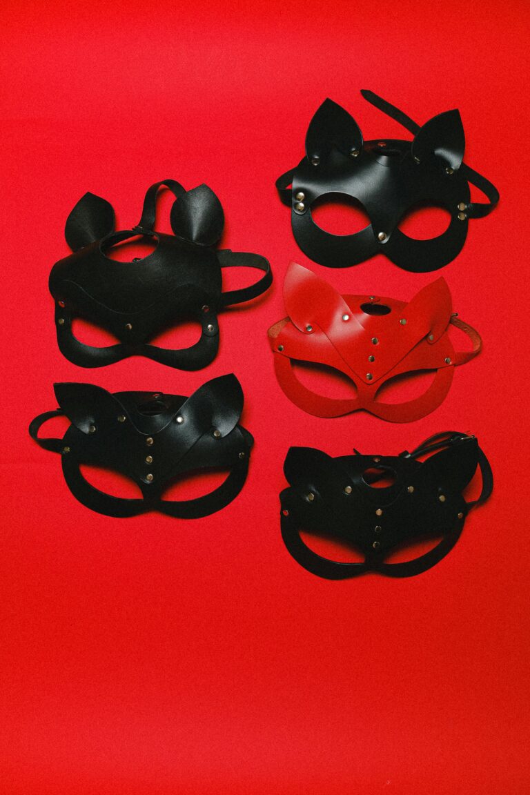 Five leather masks on a red background. Four are black, one is red. All have pointed ears, metal studs, straps, and eye and nose cutouts.