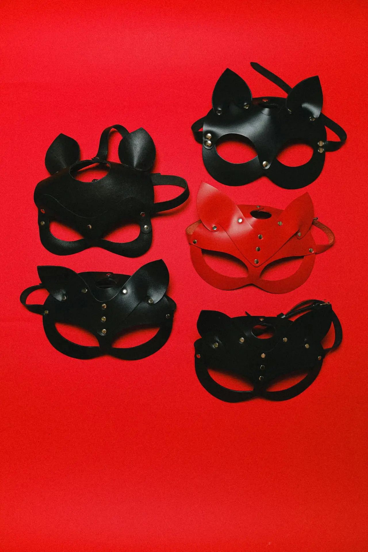 Five leather masks on a red background. Four are black, one is red. All have pointed ears, metal studs, straps, and eye and nose cutouts.