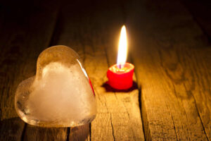A small red candle is lit next to a heart shaped ice cube on a wooden surface.