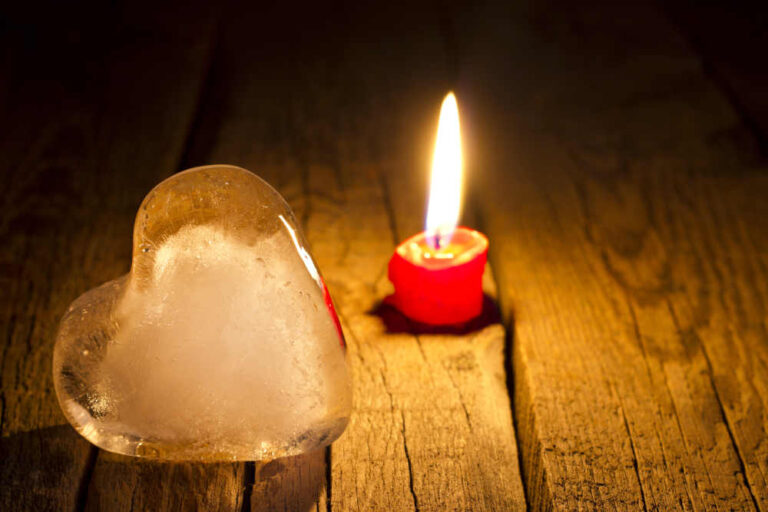 A small red candle is lit next to a heart shaped ice cube on a wooden surface.