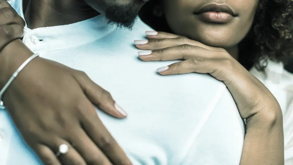 A close-up image of two people embracing, with one person's hands resting on the other's shoulders. The person in the foreground has a hand adorned with a silver bracelet and a ring, while the other person is wearing a white shirt. The background is softly blurred, focusing attention on the hands and the intimate gesture.