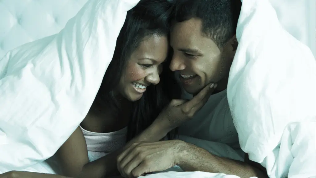 A close-up image of two people lying in bed, covered with white sheets and blankets. Both are smiling warmly at each other, with one person's hand gently touching the other's face. The background is softly lit, suggesting a cozy and intimate setting.
