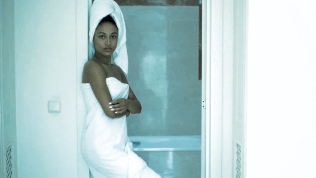 A person with a towel wrapped around their body and head stands with arms crossed in a bathroom. The individual is positioned near an open door, with a bathtub visible in the background. The bathroom features a light-colored tiled wall, a switch on the left, and a vent on the right. The overall color tone of the image is a soft blue-gray.