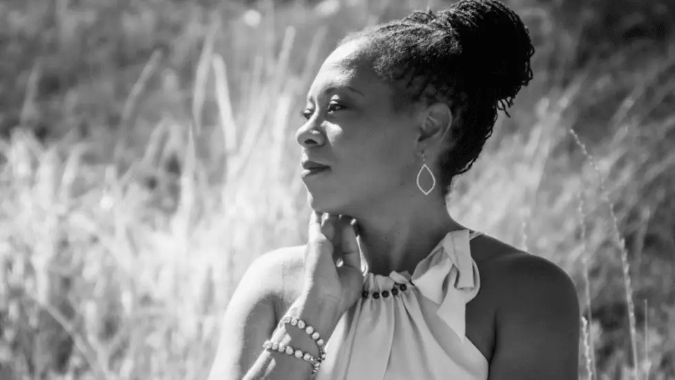 A black-and-white photograph of a woman with her hair styled in an updo, wearing dangling earrings and a sleeveless dress with a bow detail at the neckline. She is accessorized with multiple pearl bracelets on her wrist and is posing with her hand near her chin. The background features tall, blurred grass, suggesting an outdoor setting.