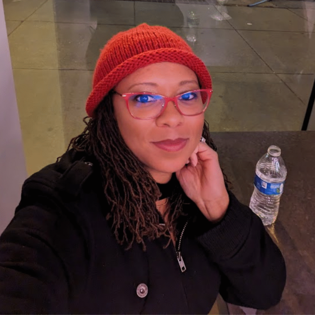 A person wearing a red knit beanie and red-framed glasses takes a selfie, with one hand resting near their face. They are dressed in a black coat with a visible zipper and button, sitting at a table with a bottle of water in the background. The setting appears to be an indoor space with a reflective floor.