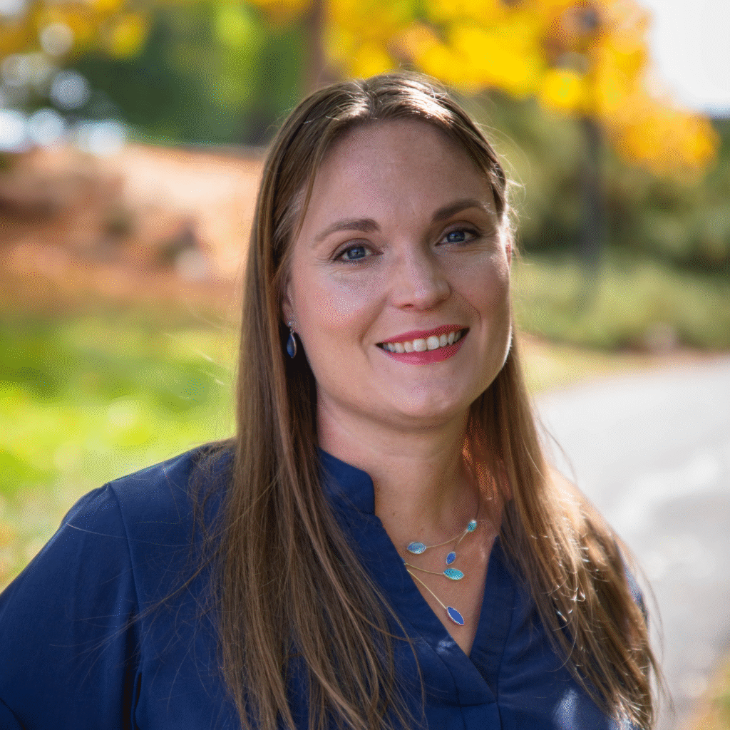 A person with long, straight brown hair stands outdoors, smiling at the camera. They are wearing a blue top and a necklace with blue and green pendant accents. The background features a scenic view with green grass, yellow autumn trees, and a pathway.