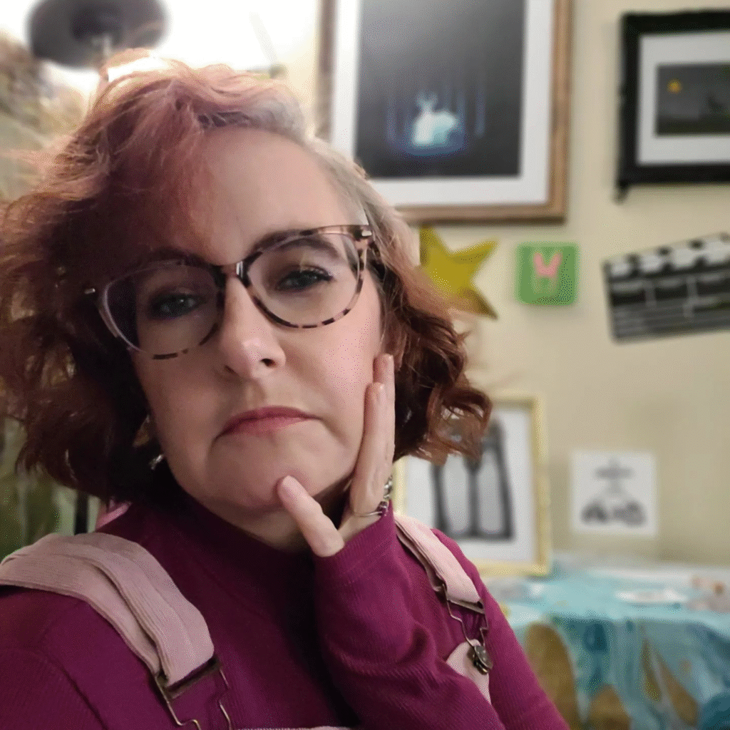 A person with curly, reddish-brown hair and glasses poses with one hand resting on their cheek. They are wearing a purple top and a pink backpack with straps over their shoulders. The background features a room with framed pictures and artwork on the walls, including a black frame with a starry image, a green star, and a clapperboard, with a table covered by a blue-green cloth in the foreground.