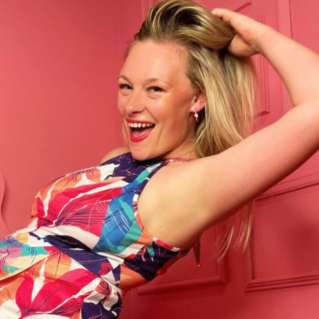 A person with long, blonde hair is captured mid-laugh, raising one arm above their head. They are wearing a colorful, floral-patterned dress with shades of pink, blue, and orange. The background is a solid pink wall with a decorative molding, suggesting an indoor setting.