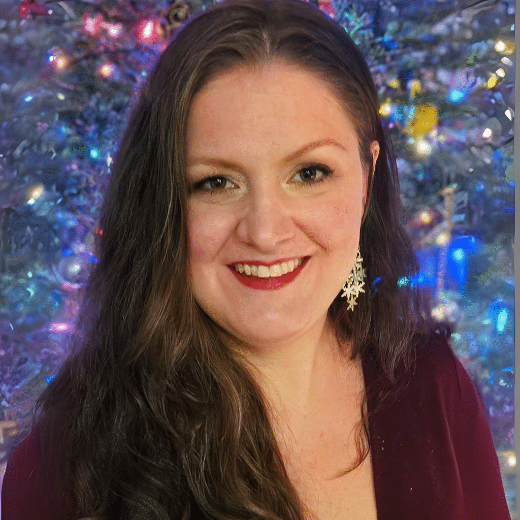 A person with long, wavy brown hair smiles at the camera, wearing a deep red top and a silver snowflake-shaped earring. The background features a festive, colorful display with blue, purple, and pink lights, resembling a decorated Christmas tree with ornaments.