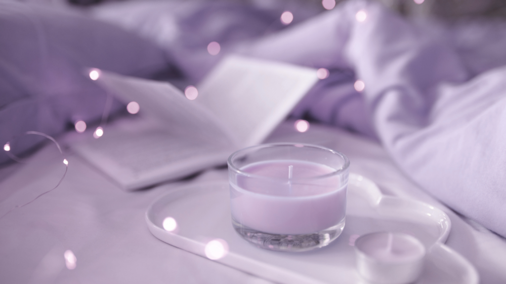 Soft purple aesthetic of a candle, open book, and fairy lights on a cozy, rumpled bed.