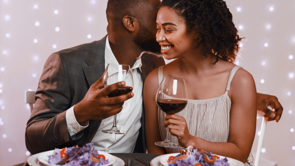 A smiling couple sharing a romantic candlelit dinner with red wine and warm string lights.
