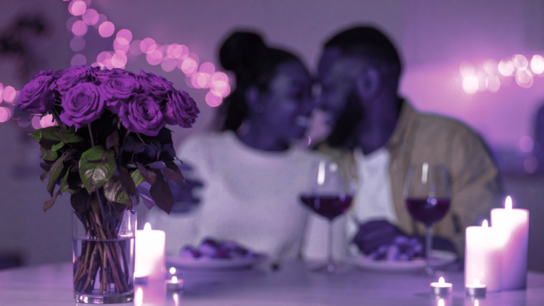 Purple-toned scene of a couple at dinner behind a bouquet of roses and glowing candles.