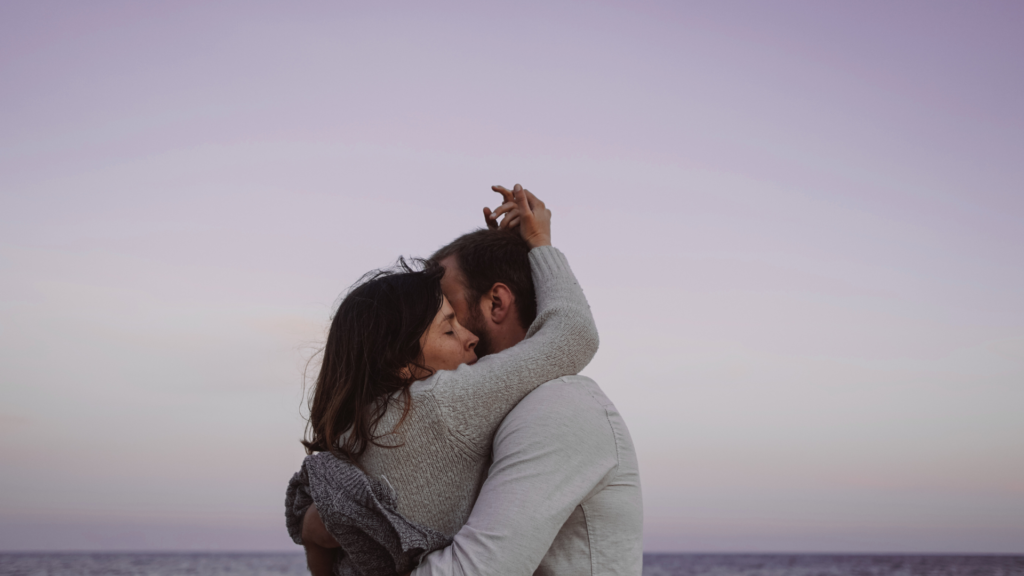 A tender embrace between a couple on a beach against a soft, twilight sky.